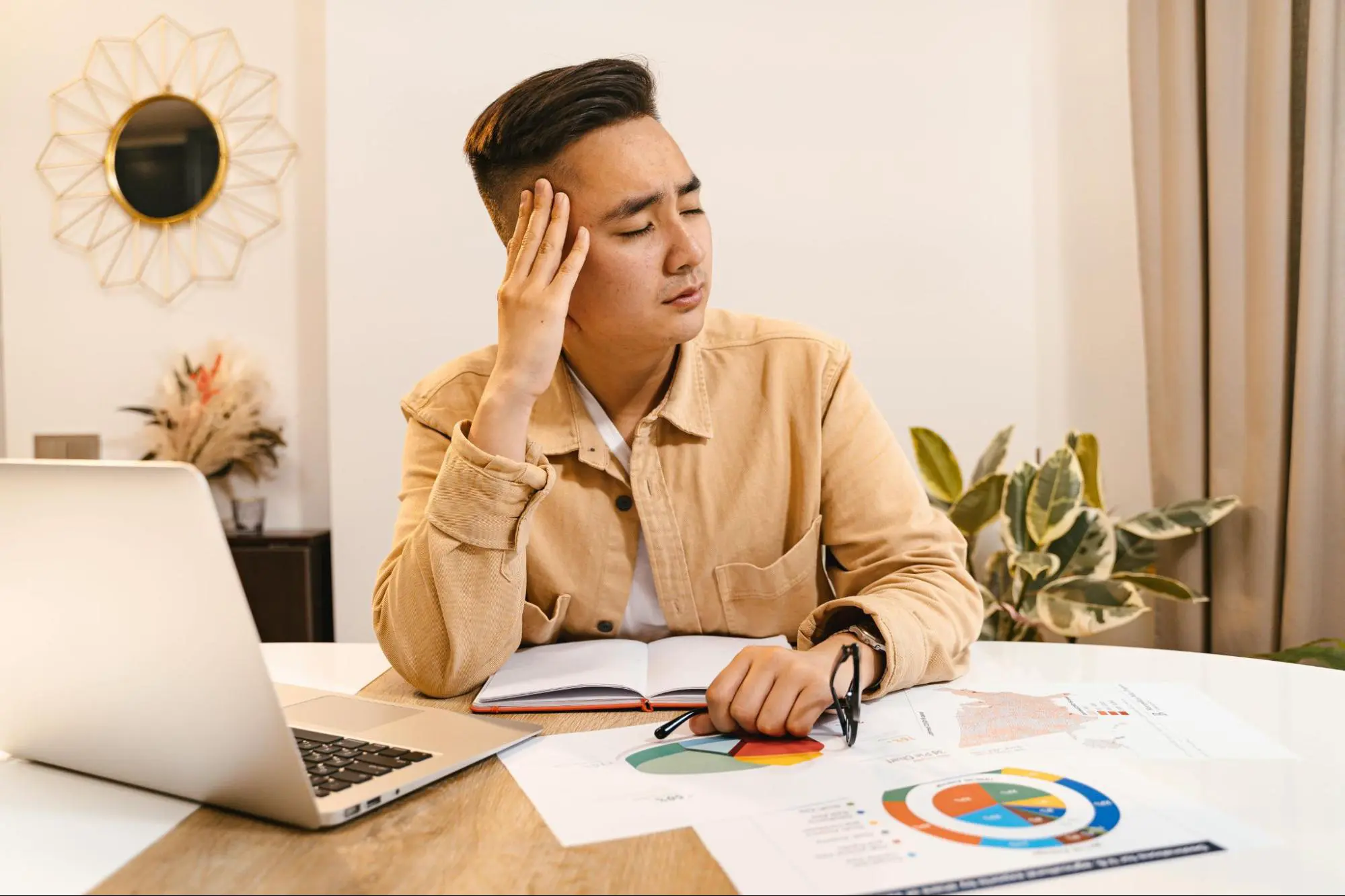 A developer at a laptop rubbing his temple, printed pie charts and market reports spread across the desk
