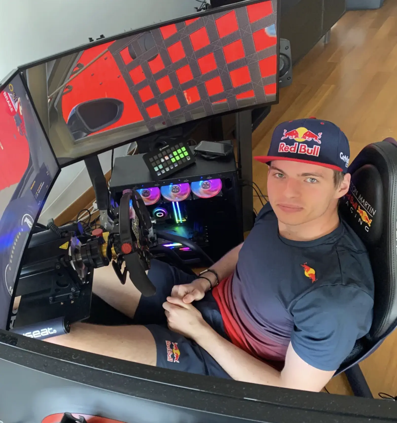 Max Verstappen in a Red Bull cap seated in a triple-screen sim racing rig with a direct drive wheel