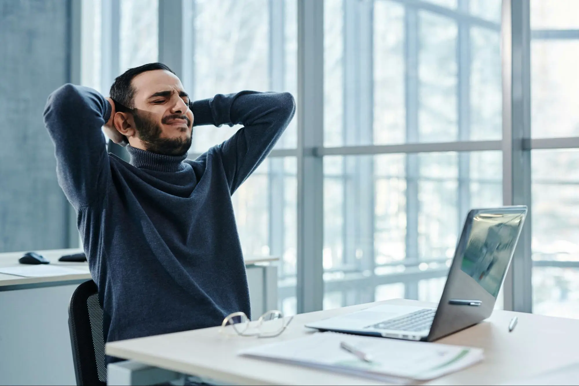 A frustrated developer leaning back with hands behind his head in front of a laptop in a bright office