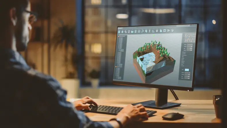 Developer at a desk building a low-poly 3D game level with trees and a water pool in a modelling tool