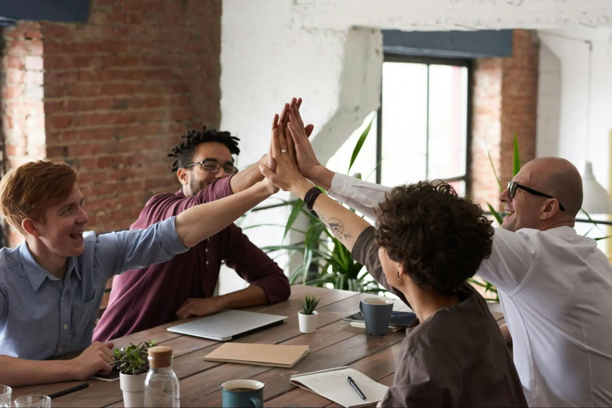 Four colleagues around a meeting table joining hands in a group high five over laptops and notebooks